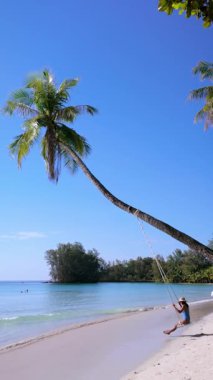 Tayland 'ın Koh Kood kentinde sakin bir günde, bir gezgin, palmiye ağaçları ve turkuaz sularıyla çevrili iken sahilde yavaşça sallanıyor. Rahatlama ve doğal güzellik bu tropikal cenneti tanımlar..