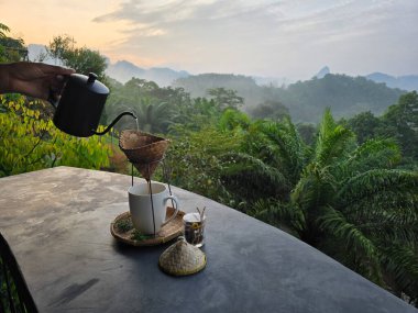 Tayland 'daki Khao Sok' un nefes kesen manzaralarını seyrederken bir fincan taze damlatılmış kahvenin tadını çıkarıyorum. Sabah sisi tropikal yeşillikleri sarar ve sakin bir atmosfer yaratır..