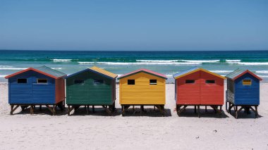 Parlak boyalı sahil kulübeleri Muizenberg sahilinin el değmemiş kumlarına karşı çarpıcı bir sahne oluşturuyor. Ziyaretçiler sıcak ve güneşli bir günün tadını çıkarıyorlar. Rahatlamak ve su içmek için mükemmel bir gün. Cape Town Güney Afrika