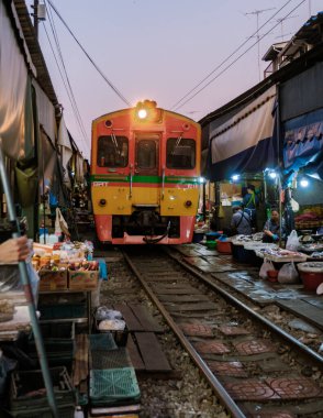 Mueang Samut Songkhram, Tayland, 4 Aralık 2025. Ziyaretçiler, alışveriş merkezlerinin ve tezgahların rayları kapladığı alacakaranlıkta canlı Maeklong Pazarı 'nda ilerleyen renkli bir trene hayret ediyor..