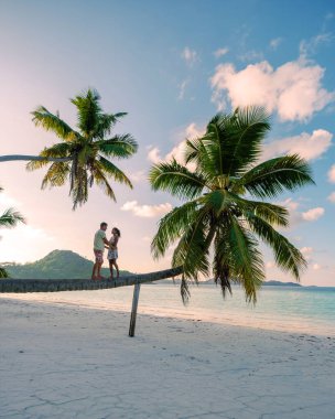 Grand Anse Sahili 'nin el değmemiş kumlarında bir çift, bir palmiye ağacında denge kurarken hassas bir anı paylaşıyor. Resimli arka planda, sallanan palmiyeler ve sakin bir okyanus manzarası yer alıyor. Praslin, Seyşeller
