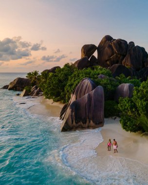 İki ziyaretçi, Anse Kaynak d 'Argent sahillerinde yüksek granit kayalar ve yemyeşil yeşilliklerle çevrili şekilde geziniyor. Göz kamaştırıcı bir günbatımı La Digue, Seyşeller 'in üzerine ılık bir ışık saçar.