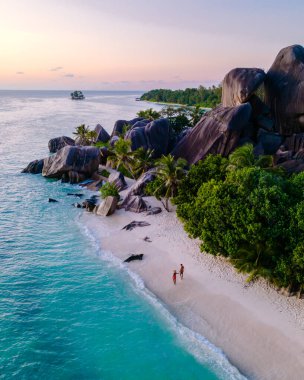 Kristal berraklığında turkuaz sular, Anse Source d 'Argent, La Digue, Seyşeller' in el değmemiş kumları boyunca yürüyen iki kişi gibi yavaşça kıyıda tur atıyor. Yükselen granit kayalar ve gür palmiyeler