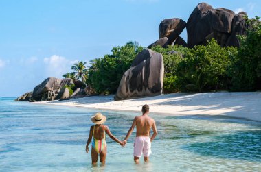 Bir çift, görkemli granit kayalar ve yemyeşil yeşilliklerle çevrili Anse Kaynak d 'Argent sahilinin sakin suları boyunca el ele yürür. La Digue, Seyşeller 'de güneşin altında mükemmel bir gün.
