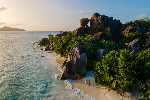 Altın güneş ışığı, Anse Source d 'Argent sahilinin ikonik granit kayalarını ve yumuşak kumlarını yıkıyor. İki gezgin, La Digue Seyşeller 'deki tropikal cenneti kucaklayarak kıyı boyunca yürür..