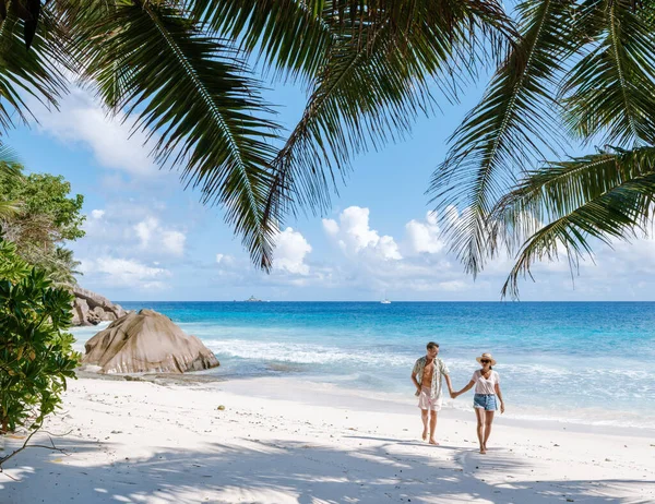 Anse Patates La Digue, Seyşeller 'de nefes kesici bir manzara, kumlu sahil boyunca el ele dolaşan bir çift, sallanan palmiyeler ve parlak mavi bir gökyüzü altında hafif dalgalarla çevrili..