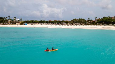 İki kanocu, Oranjestad Aruba 'daki Divi Sahili' ndeki canlı turkuaz suların keyfini çıkarıyor. Güneş, huzurlu sahilde ışıl ışıl parlıyor, rahatlatıcı bir tatil için mükemmel bir ortam yaratıyor..