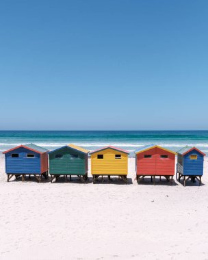 Renkli sahil kulübeleri Muizenberg sahilinin kumlu sahillerinde gururla durur, berrak mavi okyanus dalgaları dalgaları dalgalanır. Cape Town Güney Afrika