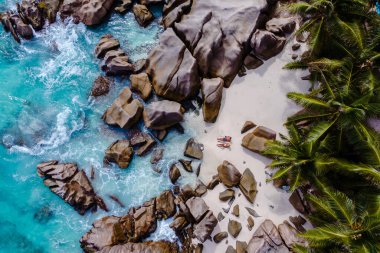 Yumuşak dalgalar Anse Patates 'teki kumlu plajda yavaşça dolaşır, güneşlenenlerin güneşin altında sakin bir günün tadını çıkardıkları yerde. Bereketli palmiye ağaçları resim çerçevesi, La Digue, Seyşeller