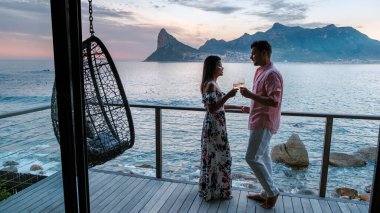 Çarpıcı sahil manzaralarının ortasında, bir çift Güney Afrika Cape Town 'da modern bir güvertede dururken içkilerimizi yudumluyor..