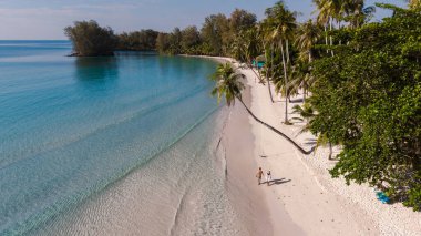 Bir çift, Tayland 'ın Koh Kood kentindeki Khlong Hin Sahili' nin yumuşak kumları boyunca hareketli palmiye ağaçları ve sakin bir turkuaz deniz ile sakin bir yürüyüş yapar..