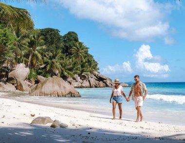 A couple enjoys a romantic walk along the pristine shores of Anse Patates in La Digue, Seychelles. The turquoise waters and lush tropical landscape create a perfect vacation backdrop.
