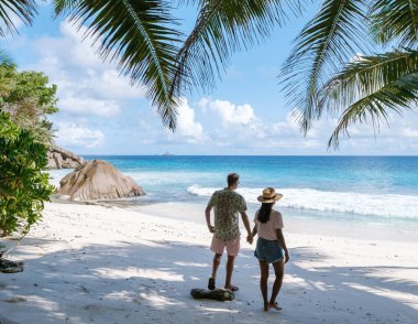 Couple enjoys a romantic walk along the tranquil shoreline of Anse Patates, surrounded by lush palm trees and crystal-clear waters. A perfect holiday getaway in Seychelles awaits.