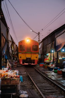 Tayland 'daki canlı Maeklong Demiryolu Pazarı' nda hareketli tezgahların ve renkli malların arasından yaklaşan bir tren görülüyor..