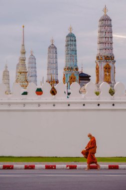 Bangkok, Tayland, 19 Kasım 2025. Güneş batarken Wat Phra Kaew 'in antik duvarlarında gezinen dingin bir keşişin manzarası. Karmaşık mimari ve ruhani atmosferi vurguluyor..