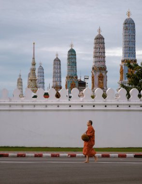 Bangkok, Tayland, 19 Novemer 2025, Bangkok sokaklarında bir keşiş bir kase tutuyor. Arka planda görkemli tapınak kuleleri yükseliyor, sakin bir sabahta zengin kültürel mirası sergiliyorlar..