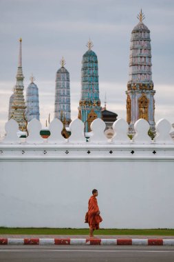 Bangkok, Tayland, 19 Kasım 2025, Bangkok 'ta parlak turuncu cübbeli bir keşiş, Wat Phra Kaeo tapınağının büyüleyici tapınak kuleleriyle çevrili bir yolda yürüyor.