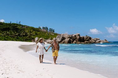 On the pristine shores of Anse Cocos beach in La Digue, Seychelles, a couple strolls hand in hand, enjoying the bright sun and beautiful turquoise waters. The scene captures romance and relaxation.