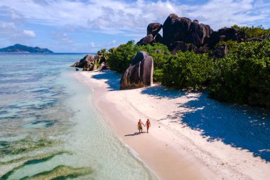 A serene beach in La Digue, Seychelles, showcases soft white sands and striking granite boulders. A couple walks hand in hand along the shore, reveling in the beauty around them during a sunny day.