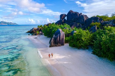 Parlak mavi bir gökyüzünün altında, Anse Kaynağı d 'Argent' ın el değmemiş kumları boyunca yürüyen iki gezgin, eşsiz granit kayalar ve yemyeşil çimlerle çerçevelenmiştir. La Digue, Seyşeller