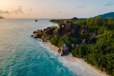 Güneş batmaya başladığında Anse Source d 'Argent' ın el değmemiş sahil şeridine hafif dalgalar vurur. Sahne, çarpıcı granit kayalar ve yemyeşil, La Digue, Seyşeller ile dolu.