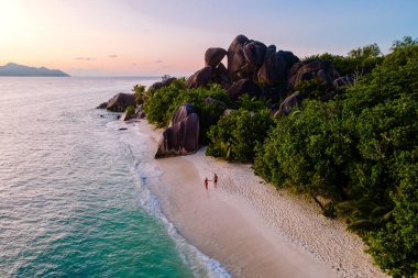 Dalgalar yavaşça Anse Kaynağı d 'Argent kıyılarını dolaşırken iki insan el değmemiş kumlarda geziniyor, güneş batarken görkemli kayalar ve yemyeşil kayalarla çevrili, La Digue, Seyşeller