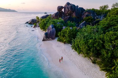 Ziyaretçiler gün batımında Anse Source d 'Argent plajında yürürler. Yükselen granit kayalar ve yemyeşil alanlar Seyşeller 'de sakin bir tropik kaçış yaratır..