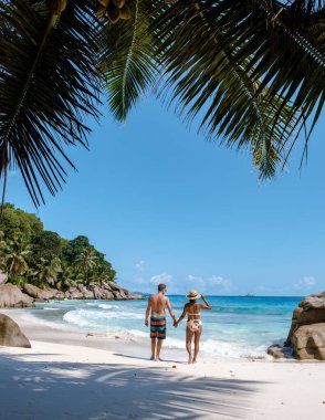 A couple strolls hand in hand along the pristine shoreline of Anse Patates, La Digue, Seychelles. Surrounded by lush greenery and azure waters, the scene captures a perfect tropical escape.