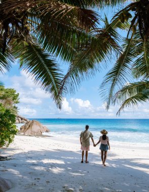 Bir çift el ele yürür Anse Patates 'in el değmemiş kıyıları boyunca, yemyeşil palmiye ağaçları ve canlı mavi okyanuslarla çevrili. Güneş parlıyor, La Digue, Seyşeller