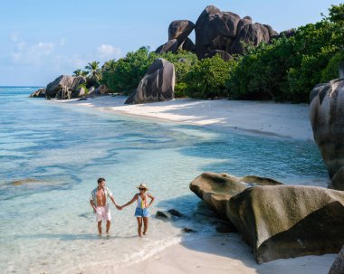 A couple enjoys a tranquil afternoon walk hand in hand at Anse Source d'Argent, surrounded by stunning rock formations, lush greenery, and crystal-clear waters of La Digue Seychelles.