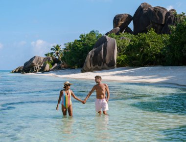 Crystal clear waters gently lap at the shores of Anse Source d'Argent as a couple walks hand in hand. La Digue, Seychelles