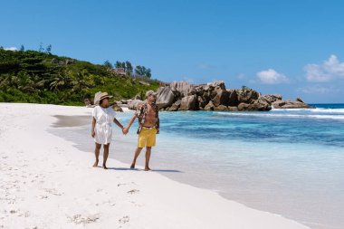 Soft white sands stretch along Anse Cocos beach as a couple strolls hand in hand, surrounded by stunning turquoise waters and lush greenery under a clear blue sky. La Digue Seychelles