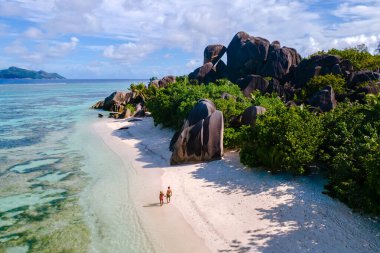 Two travelers walk slowly along the pristine beach of Anse Source d'Argent, surrounded by towering granite rocks of La Digue, Seychelles. The vibrant turquoise waters reflect a perfect tropical day.