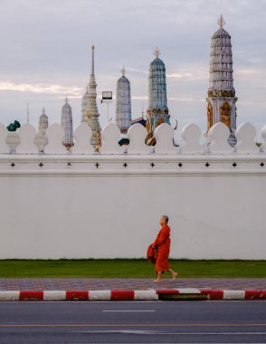 Bangkok, Tayland, 19 Kasım 2025 'te Wat Phra Kaew' in beyaz duvarı ve karmaşık kuleleri tarafından çerçevelenmiş turuncu cüppeli bir keşişi sokakta huzur içinde yürürken yakalıyor..