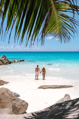 A couple walks hand in hand along the stunning white sand beach of Anse Georgette in Praslin, Seychelles. The vibrant turquoise sea sparkles under the warm sun, creating a perfect tropical escape.
