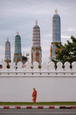 Bangkok, Tayland, 19 Kasım 2025, geleneksel turuncu cübbe giymiş bir keşiş, Wat Phra kaeo 'nun karmaşık desenleriyle süslü, çarpıcı tapınak kulelerinin önünden geçiyor..