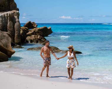 A couple walks hand in hand along the pristine sands of Anse Cocos beach, embraced by turquoise waters and majestic rock formations. La Digue Seychelles