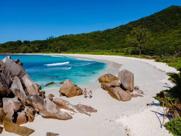 Yumuşak beyaz kumların berrak turkuaz sularla buluştuğu Anse Cocos plajının huzurlu güzelliğinin tadını çıkarın. Bir çift kadın ve erkek kumsalda yürüyor, İHA hava görüntüsü La Digue Seyşeller