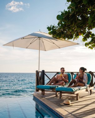 İki arkadaş, Willemstad, Curacao 'da sakin bir tatil köyünde uzanırken içkileri tazelerken keyif çatıyor. Altın güneş ışığı, gün batımında havuz kenarında rahatlatıcı bir tatil için mükemmel bir zemin çizer.