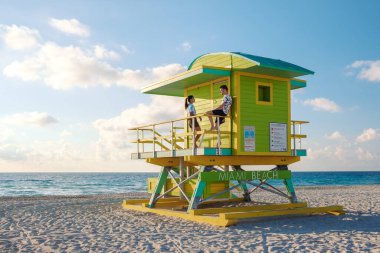 Parlak sarı cankurtaran kulesi Miami Plajı 'nı gözlüyor. İnsanlar sıcak kumda dinleniyor. Açık mavi gökyüzünün altında büyüleyici sahil manzarasının ve canlı atmosferin tadını çıkarıyorlar..