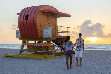 Miami Sahili 'nin kumlu sahillerinde, nefes kesen günbatımından büyülenmiş bir çift el ele yürüyor. Canlı cankurtaran duruşu bu dingin plaj anına renkli bir dokunuş katıyor..