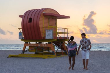 Güneş Miami sahilinin üzerinde batarken, bir çift renkli bir cankurtaran kulübesinin yanında el ele yürüyor. Sakin atmosfer romantik bir tatilin özünü yakalar..