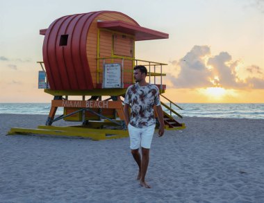 Güneş batarken Miami sahilinin yumuşak kumlarında yürüyen bir adam, sıcak altın renklerini gökyüzüne saçıyor ve yanındaki renkli cankurtaran kulesini aydınlatıyor..