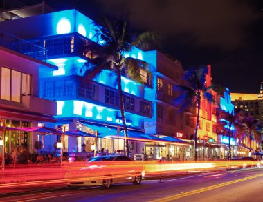 Miami Plajı 'ndaki Ocean Drive boyunca canlı ışıklar sanat deco binaları boyunca dans ediyor. Ziyaretçiler gece hayatının tadını çıkarırken arabalar geçiyor ve heyecan dolu dinamik bir atmosfer yaratıyorlar..