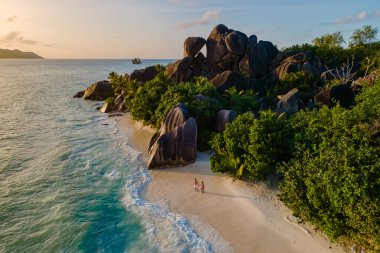 La Digue, Seyşeller 'deki Anse Source d' Argent plajı boyunca yürüyen iki gezgin, sakin bir günbatımının altında, granit kayalar ve yemyeşil yeşilliklerle çevrili, hafif dalgalar kıyıları öpüyor..
