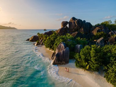 Çiftler Anse Source d 'Argent' ın beyaz kumlu plajında yürürken yumuşak dalgalar sahili okşar. La Digue, Seyşeller 'de kumsalda bir çift yürüyor, insansız hava aracı manzaralı.