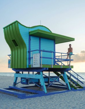 Miami Sahili 'ndeki çarpıcı bir cankurtaran kulesi gün batımına karşı koyu mavi ve yeşil renkler sergiliyor. Rahatlamış bir adam, sakin manzaranın tadını çıkarırken, tatil ruhunu somutlaştırıyor..