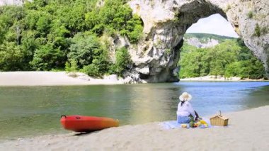 Fransa 'nın Ardeche kentindeki Pont dArc nehrinde huzurlu bir günün tadını çıkarın. Parlak bir kano yakınlarda dinlenirken, bir kadın piknik yaparken dinleniyor, yemyeşil ve kayalık kayalıklardan oluşan sakin bir zemin sunuyor..