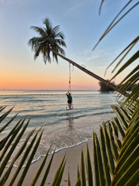 Alacakaranlıkta Ko Kut 'un yumuşak kumlu plajında büyüleyici bir sahne başlıyor. Yeşilliğin çerçevelediği eğimli bir palmiye ağacında salıncaktan hoşlanan bir insan..