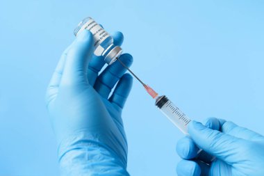 Mavi eldivenli bir doktorun elleri mavi arka planda aşı için şırıngayı hazırlıyor.